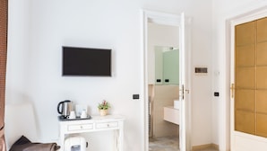 A hotel room with a white desk, a television, a coffee maker, a water dispenser, a small plant, and a bathroom visible through an open door. - Relais Calamatta (Rome)