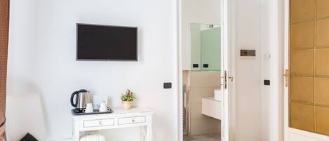 A hotel room with a white desk, a television, a coffee maker, a water dispenser, a small plant, and a bathroom visible through an open door.