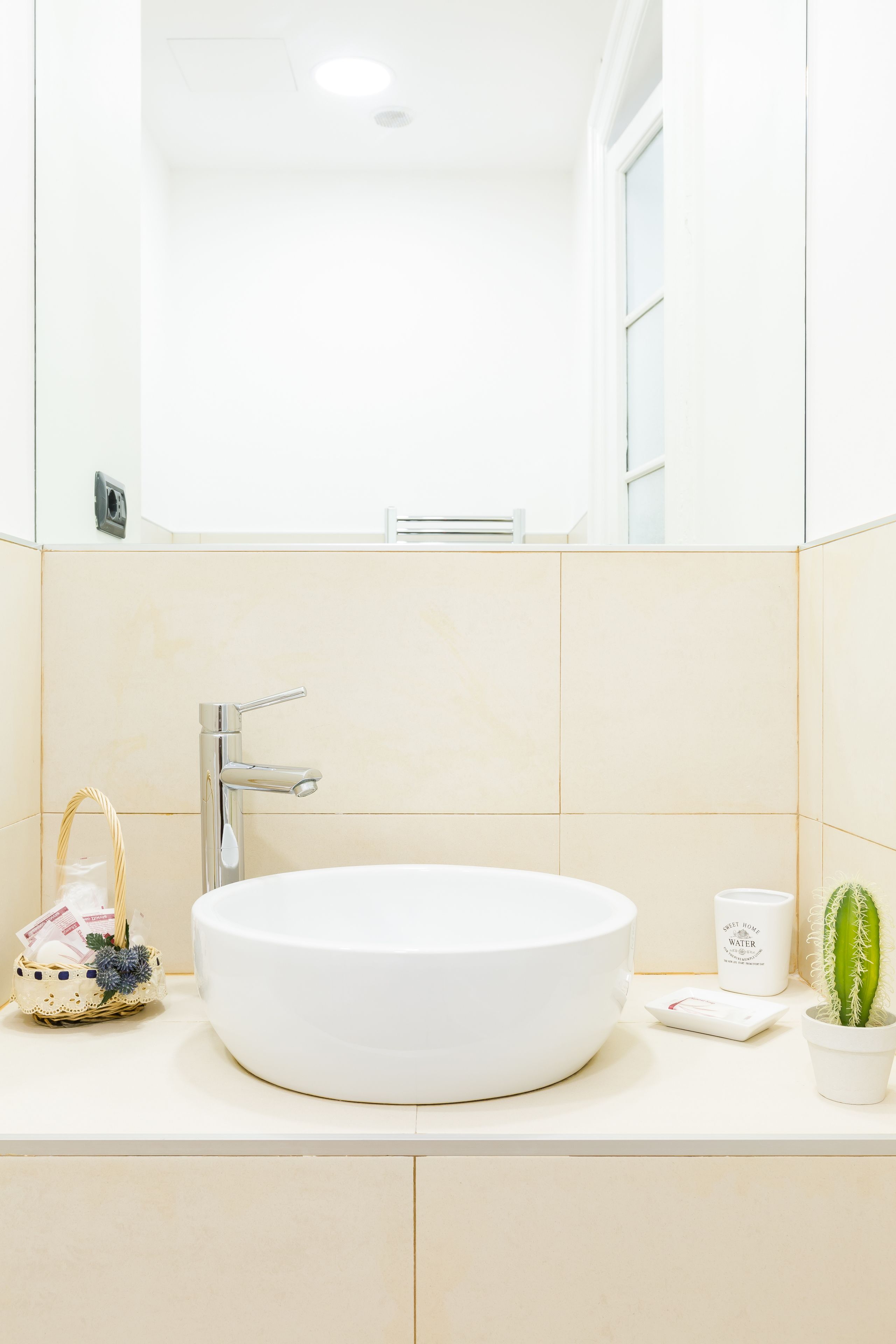 A bathroom with a round white sink, a chrome faucet, a small wicker basket with a bag, a white candle, and a potted cactus.