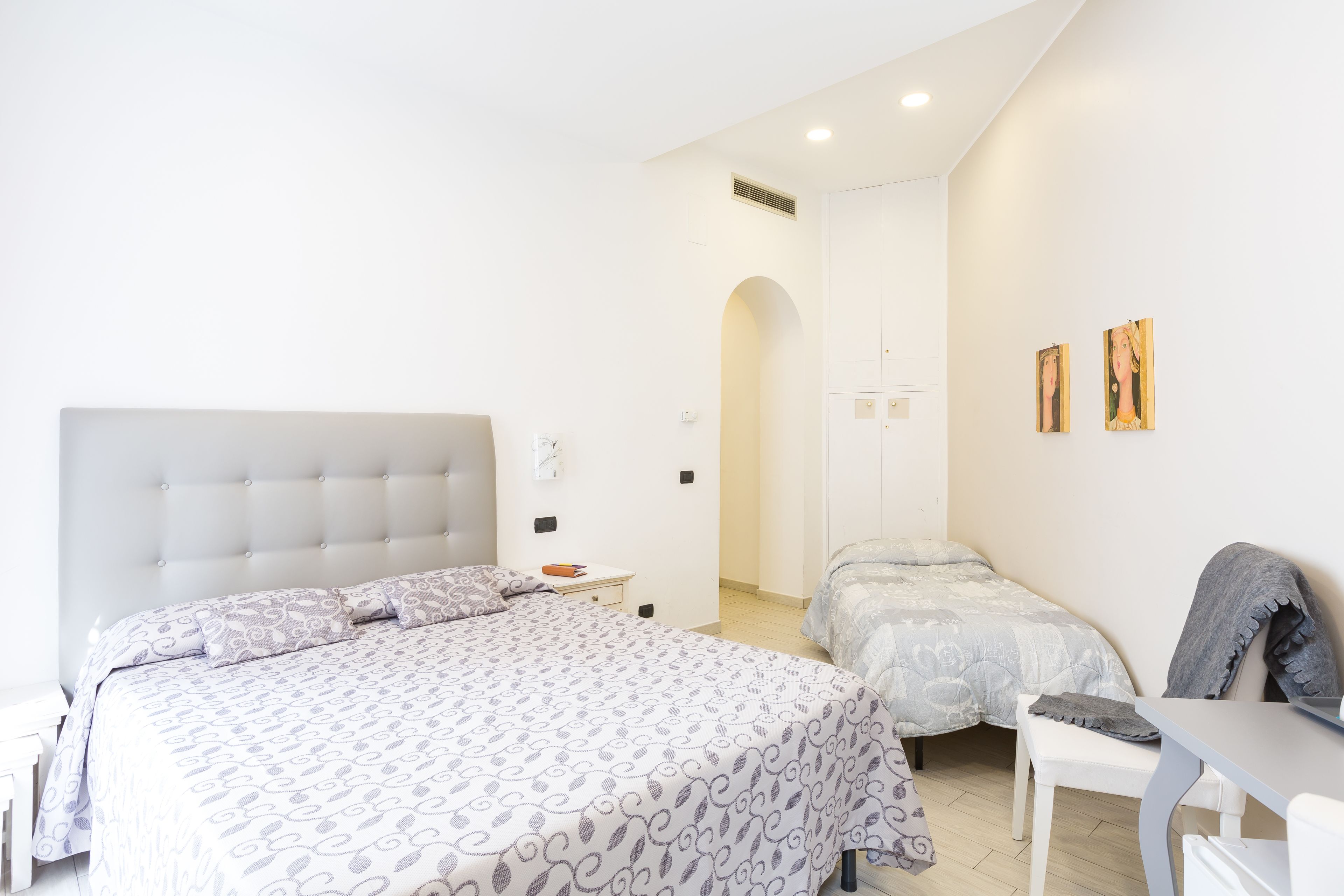 A bedroom with a large bed, a smaller bed, a desk, and a chair. There is a white wall with two paintings.