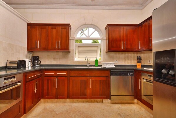 A kitchen with wooden cabinets, stainless steel appliances, a window with blinds, and a tiled backsplash.
