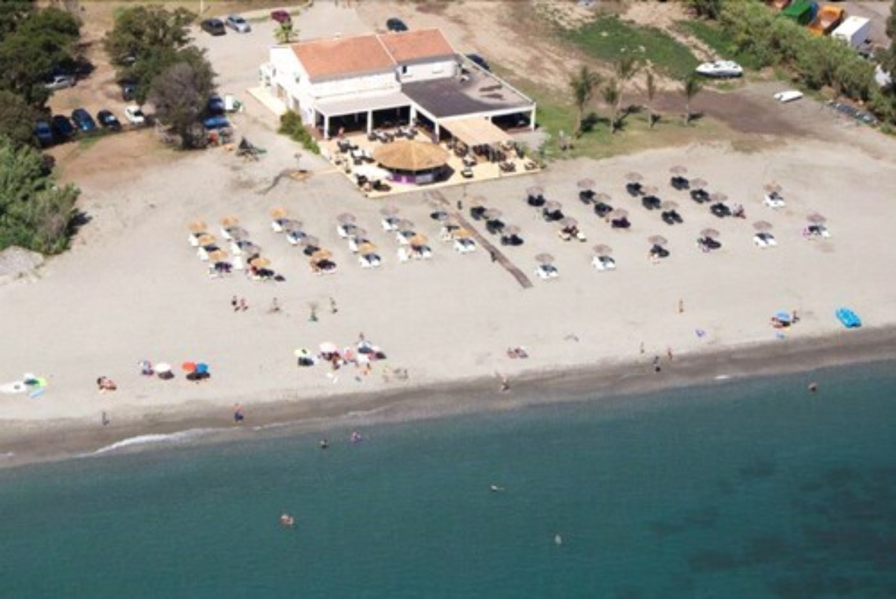 Vista aérea de una propiedad frente a la playa con un edificio grande, varios espacios de estacionamiento y una zona de arena.