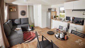 A modern living room with a grey sectional sofa, a small round table, and a dining area with a wooden table and chairs. - Résidence Marea Resort (Santa-Lucia-di-Moriani)