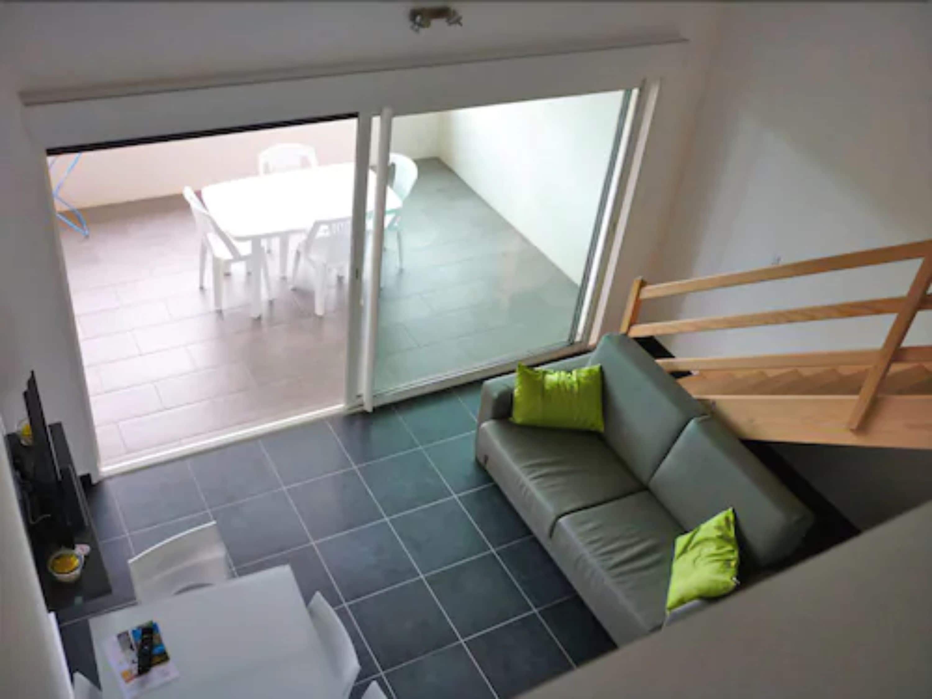 Un salon moderne avec un canapé gris, des coussins verts, une table basse blanche et une porte-fenêtre coulissante donnant sur un balcon aménagé en coin repas.