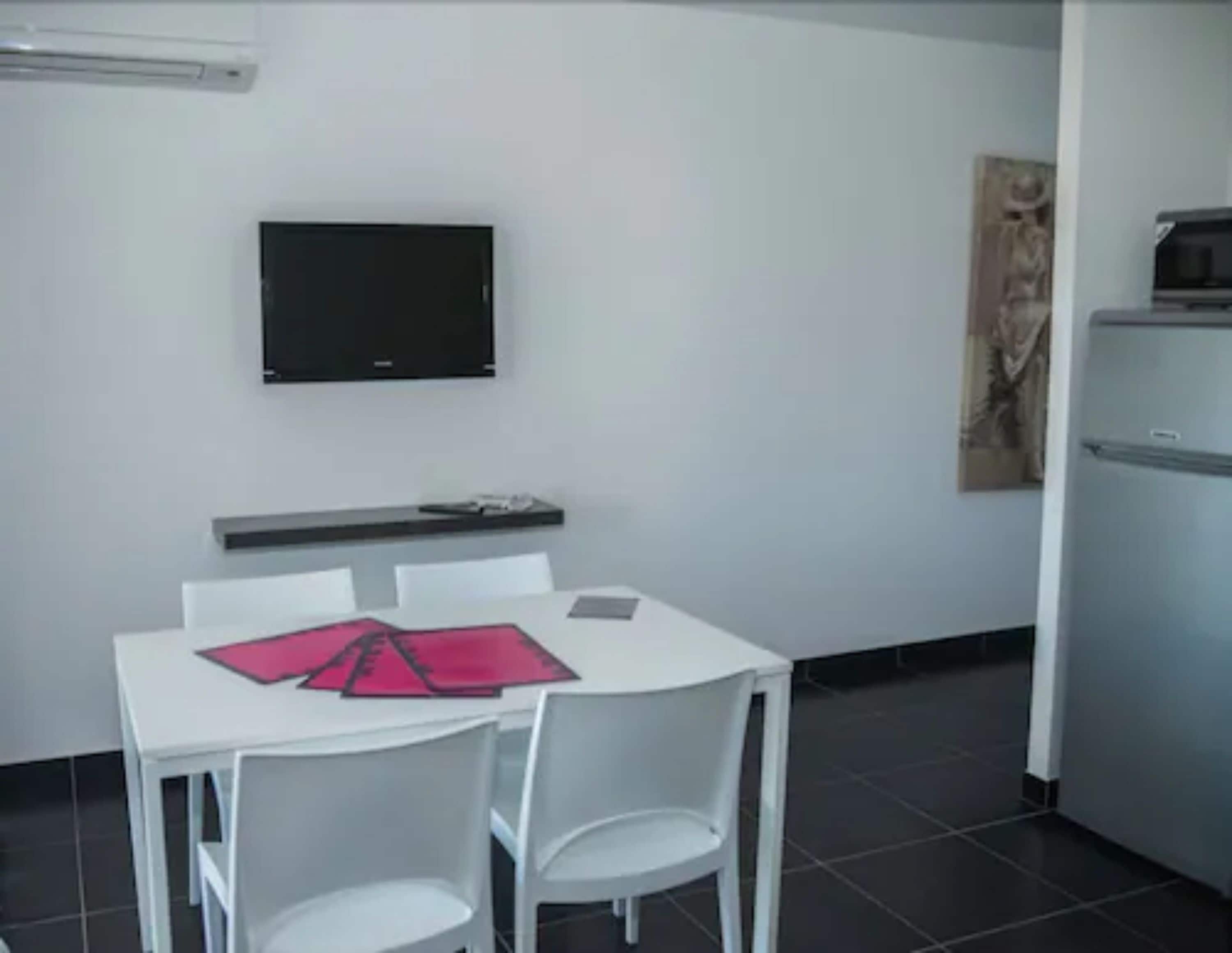 A small, modern room with a white table and chairs, a flat-screen TV mounted on the wall, a refrigerator, and a tiled floor.