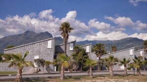 A modern building with a stone facade, surrounded by palm trees and mountains in the background. - Résidence Marea Resort (Santa-Lucia-di-Moriani)