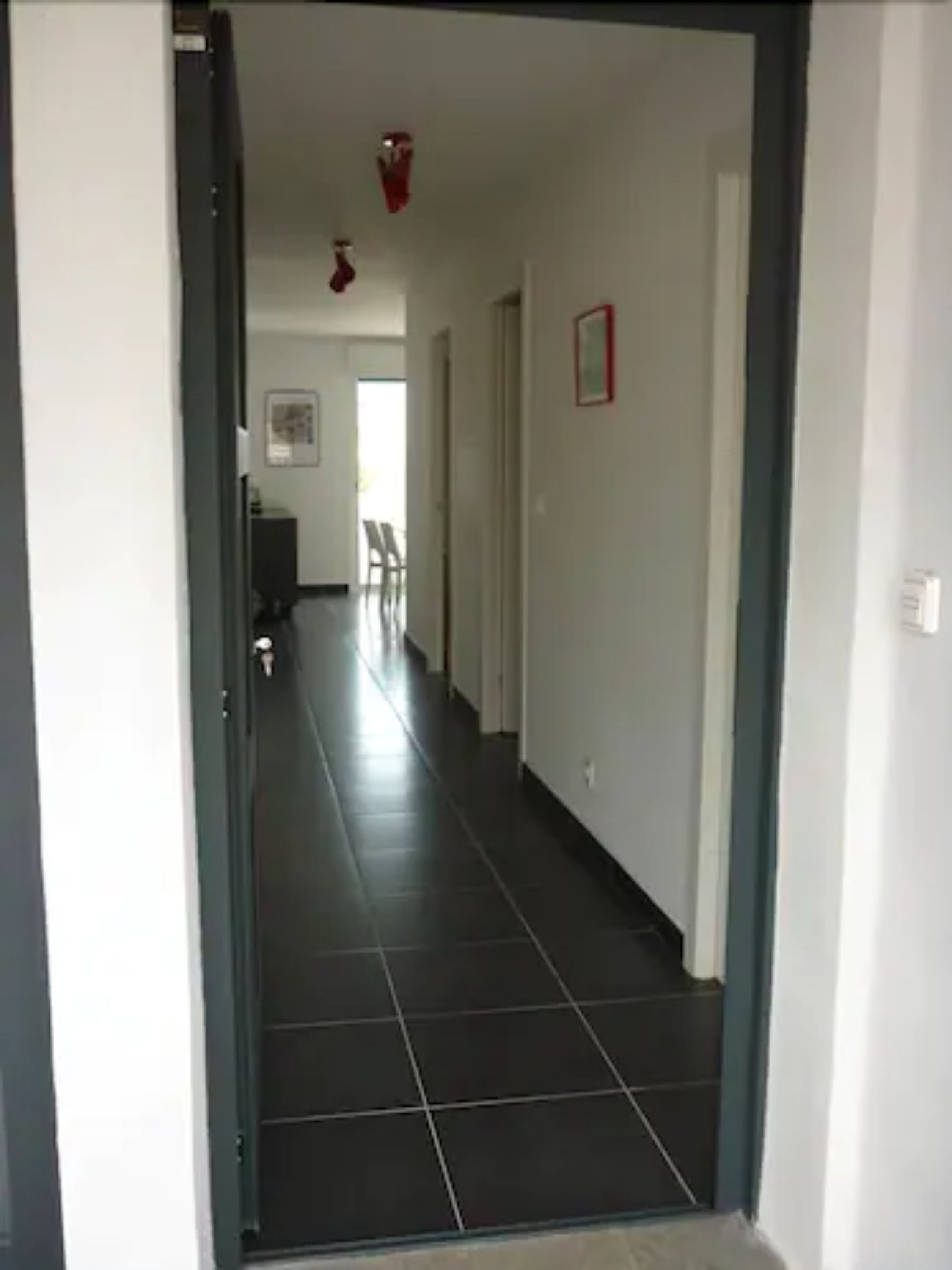 Un couloir avec un sol carrelé foncé, des murs blancs et une porte sur le côté gauche.