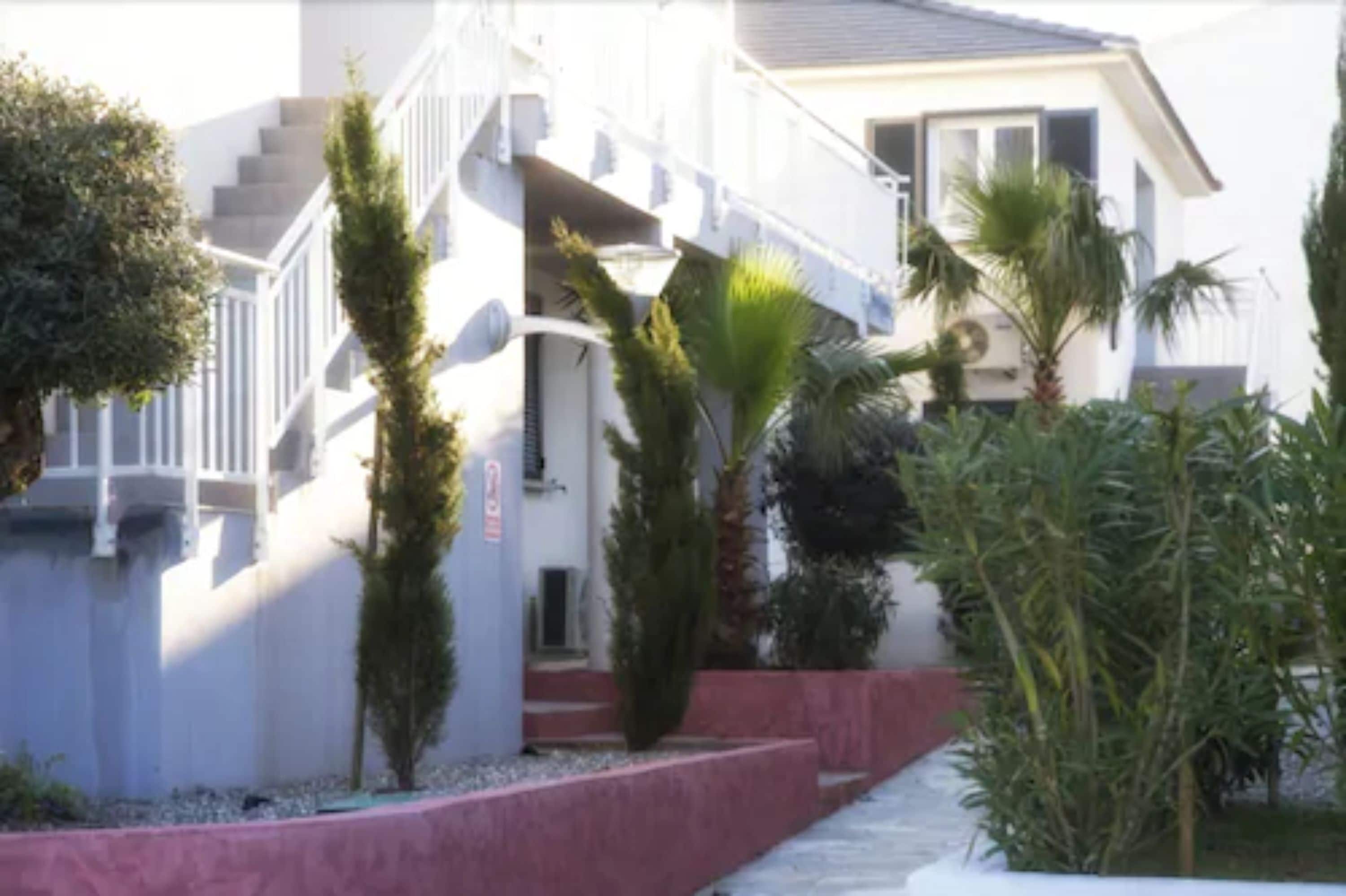 A two-story building with a staircase leading to the upper floor. There are palm trees and other greenery in the front yard.