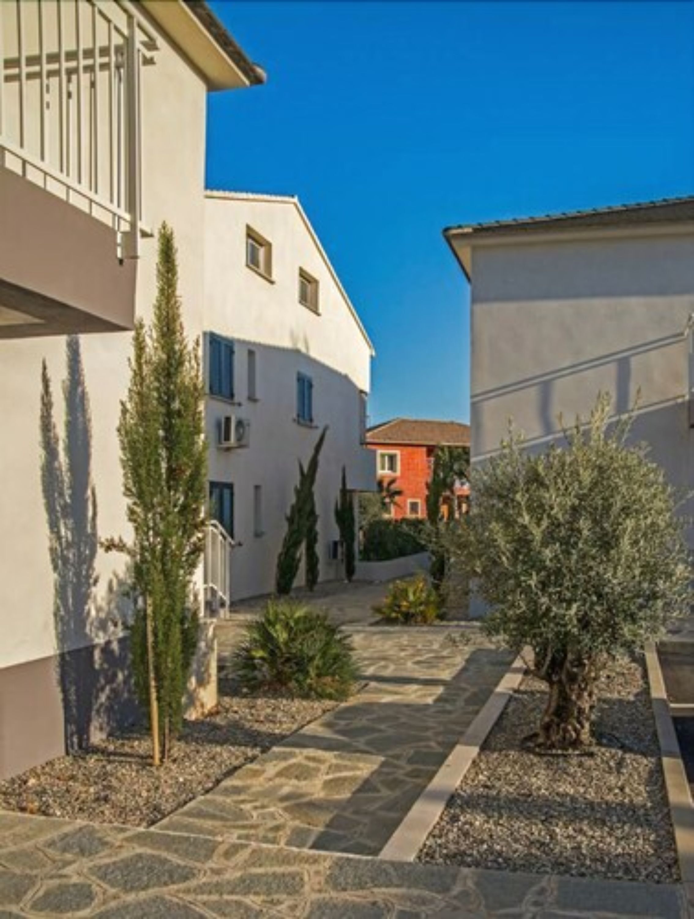 A courtyard with stone pathways, a small tree, and a building with a white facade and blue shutters.