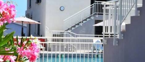 Un área de piscina con valla blanca, una escalera que conduce a un edificio y flores rosadas en primer plano.