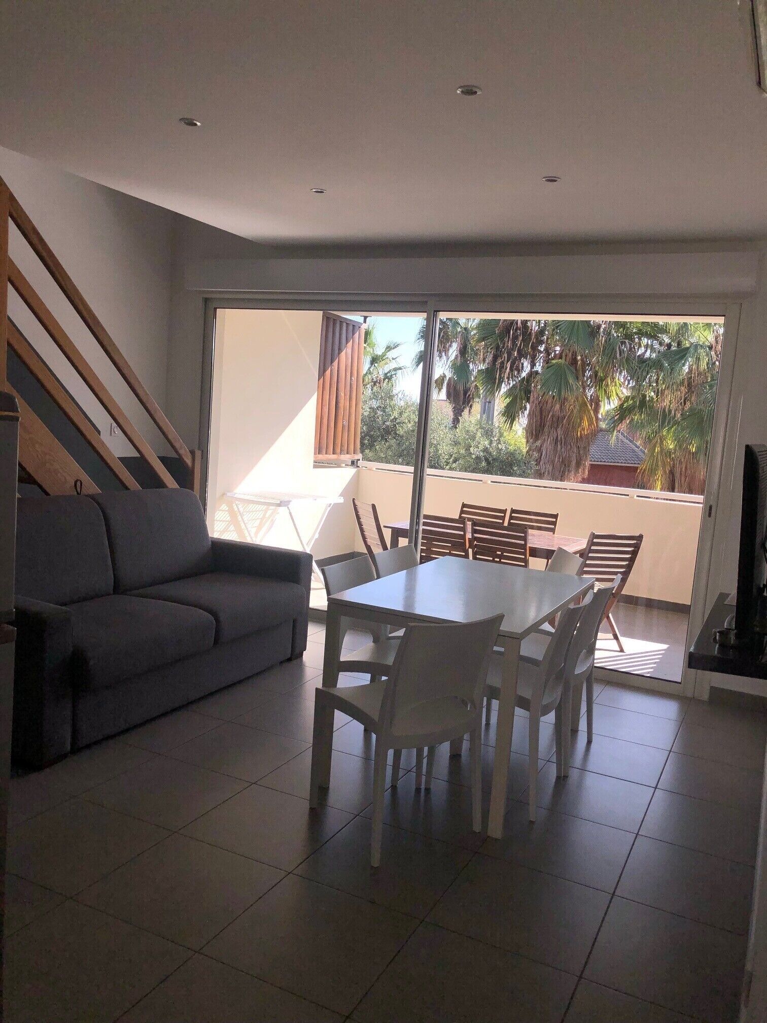 A modern living room with a grey sofa, a dining table with white chairs, and a large sliding glass door leading to an outdoor balcony with palm trees.