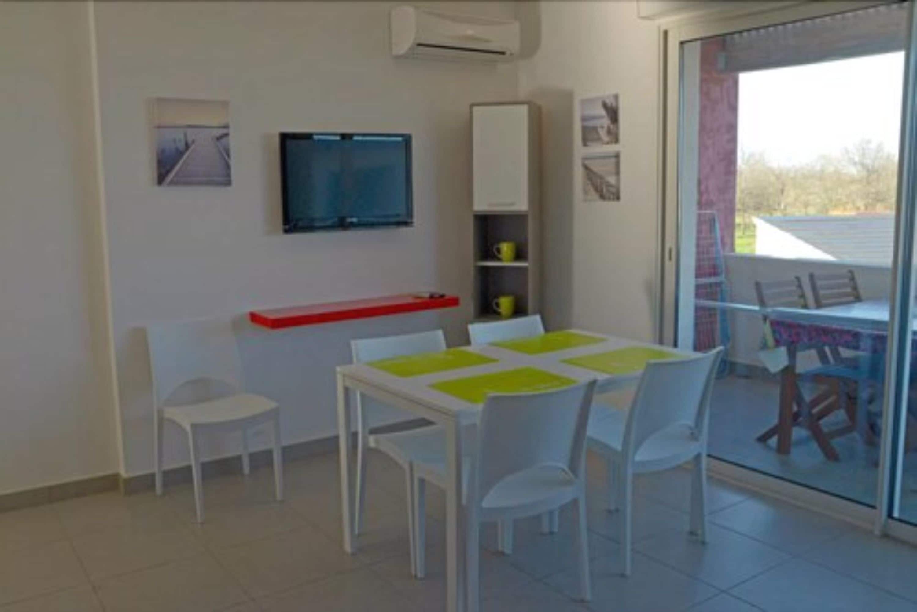 Zona pranzo con tavolo e sedie, televisione a parete e porta scorrevole in vetro che dà su un'area esterna.