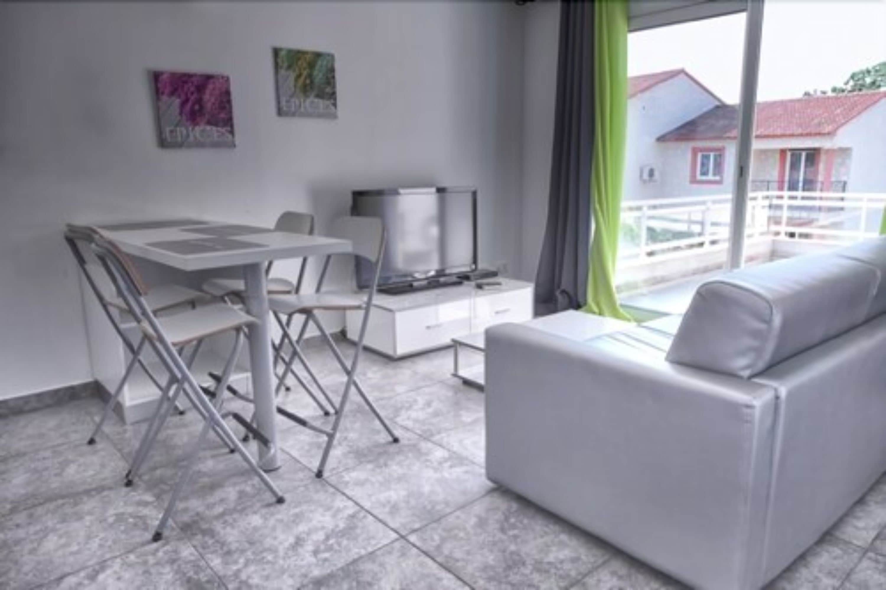 A modern living room with a white sofa, a small table, and a TV. There are two paintings on the wall and a balcony with a railing.
