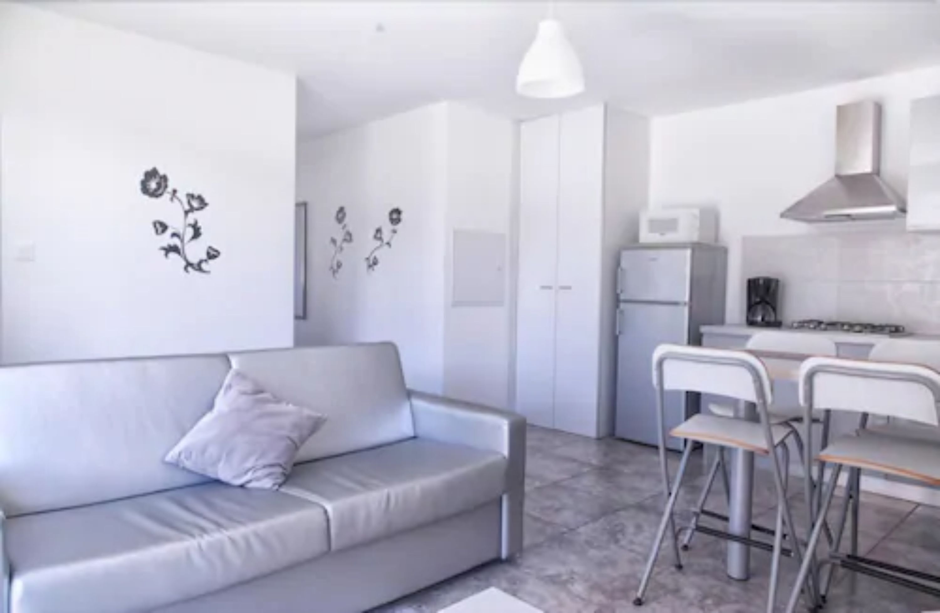 A modern kitchen with white cabinets, a stove, and a microwave. There is a grey sofa with a pillow and two metal chairs.