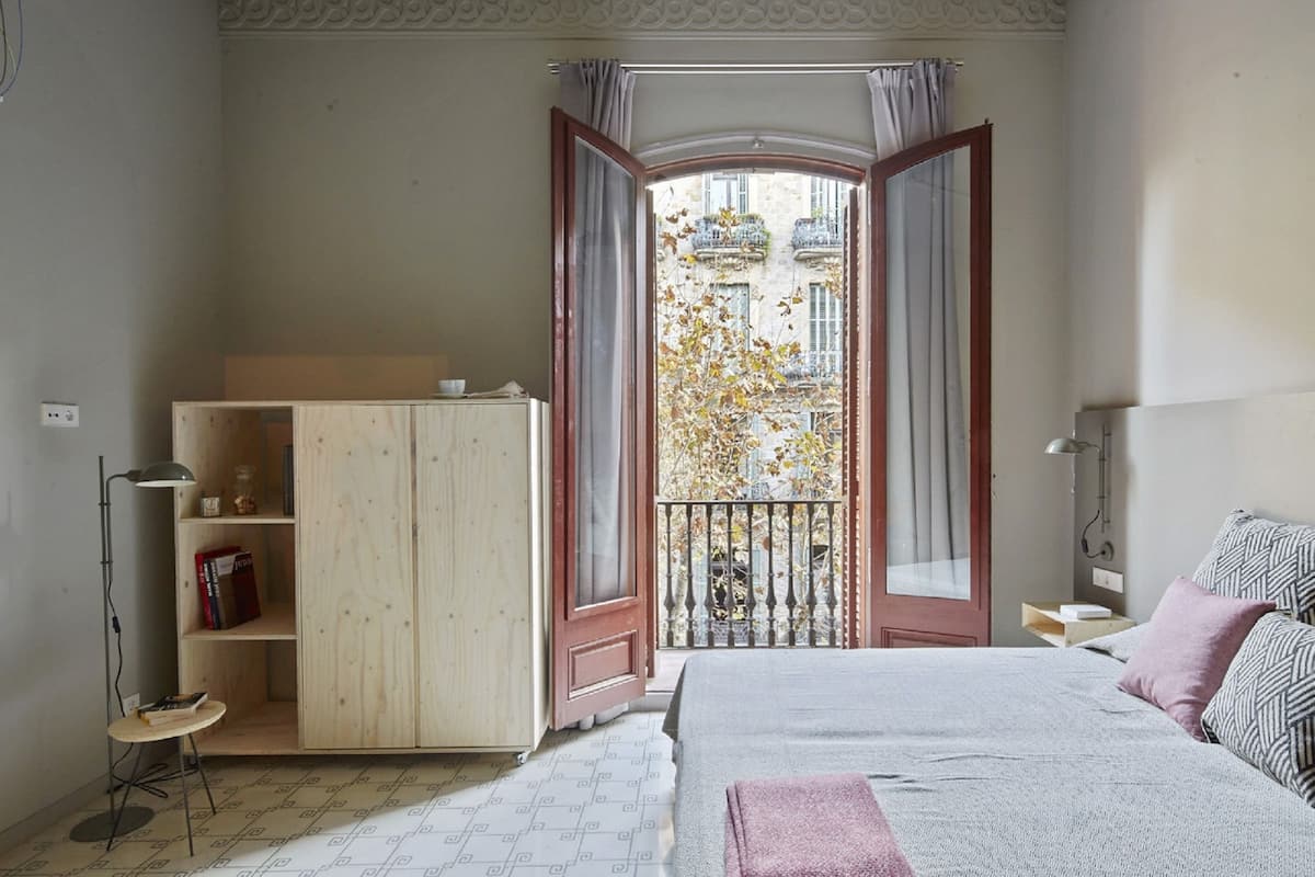 Een slaapkamer met een bed, een houten garderobe, een klein tafeltje en een balkon met uitzicht op gebouwen.