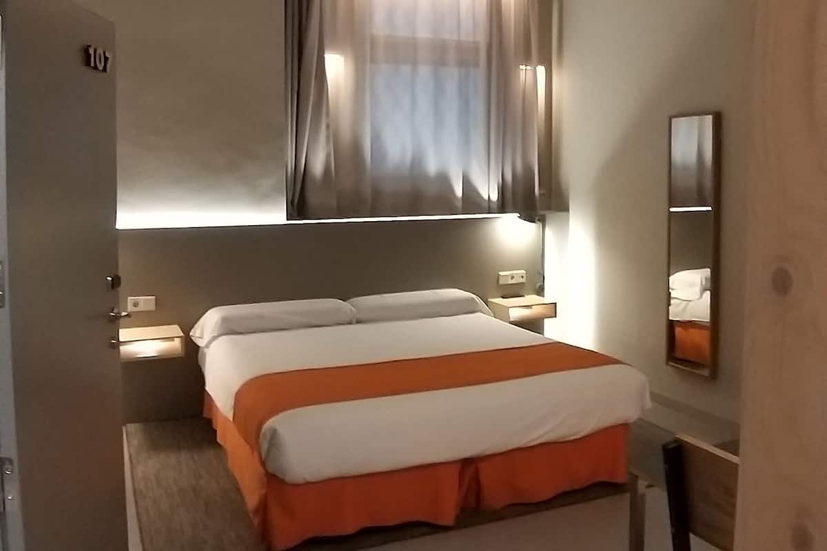 Een slaapkamer met een bed, een raam met gordijnen, een nachtkastje en een spiegel.