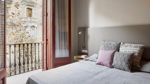 A bedroom with a bed, pillows, a nightstand, a lamp, and a view of a building with balconies and trees. - Casa Abamita (Barcelona)