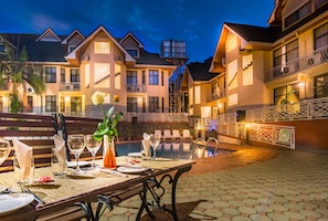 Un área de comedor al aire libre bien iluminada con piscina, mesas y sillas para comer, rodeada por un edificio grande con múltiples balcones y una fuente.