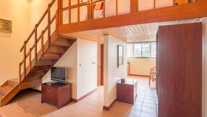 Een ruime woonkamer met een houten trap, een flatscreen televisie op een houten standaard, en een eethoek met stoelen.