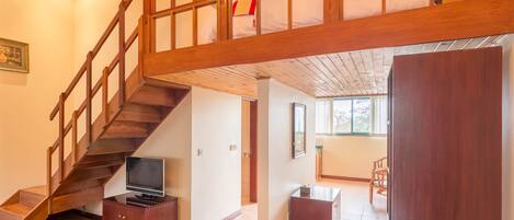 A spacious living area with a wooden staircase, a flat-screen TV on a wooden stand, and a dining area with chairs.