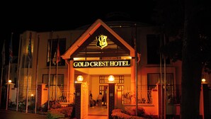 Het exterieur van het Gold Crest Hotel 's nachts, met een opvallende ingang, een bord, een goed verlichte gevel en een aangelegde tuin.
