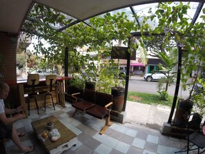 Uma área externa com mesas e cadeiras, cercada por plantas em vasos e vista para a rua.