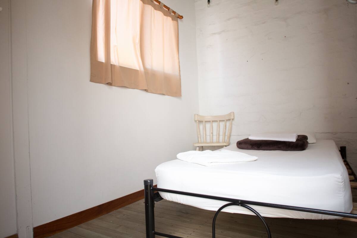 Un lit simple avec un matelas blanc et une tête de lit, une chaise en bois et une fenêtre avec un rideau beige.