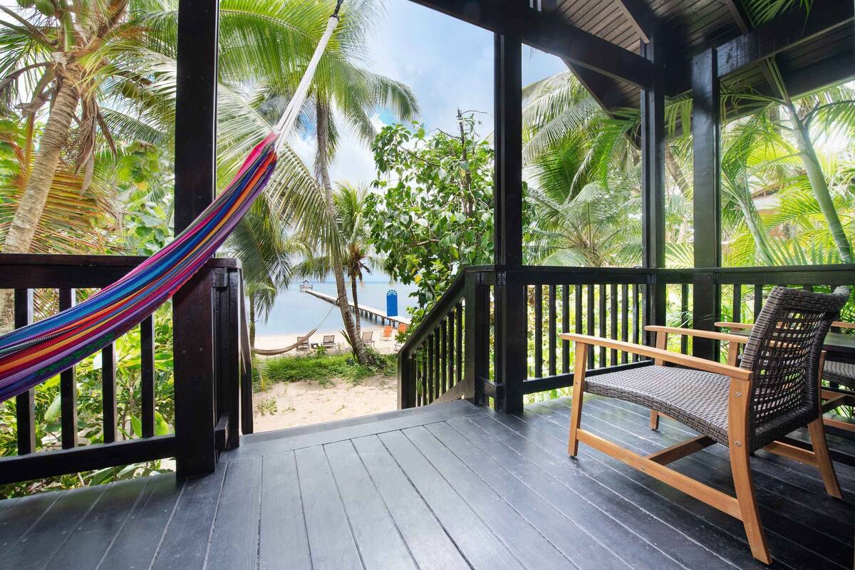 A wooden deck with a hammock and wicker furniture, overlooking a beach and palm trees.