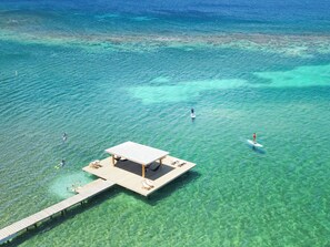 An aerial view of a clear blue sea with a wooden pier leading to a platform with a roof, surrounded by clear water and a few people paddleboarding.