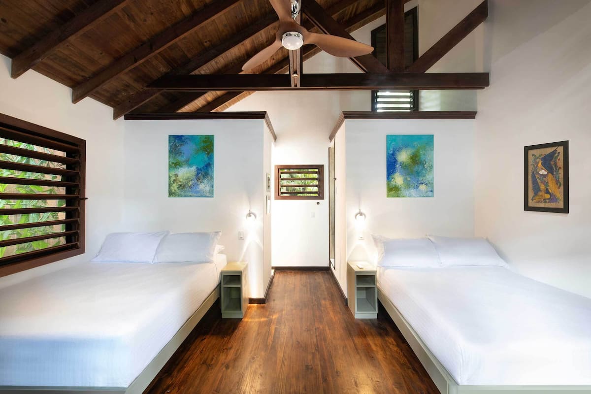 A room with two beds, wooden flooring, a ceiling fan, and artwork on the walls.