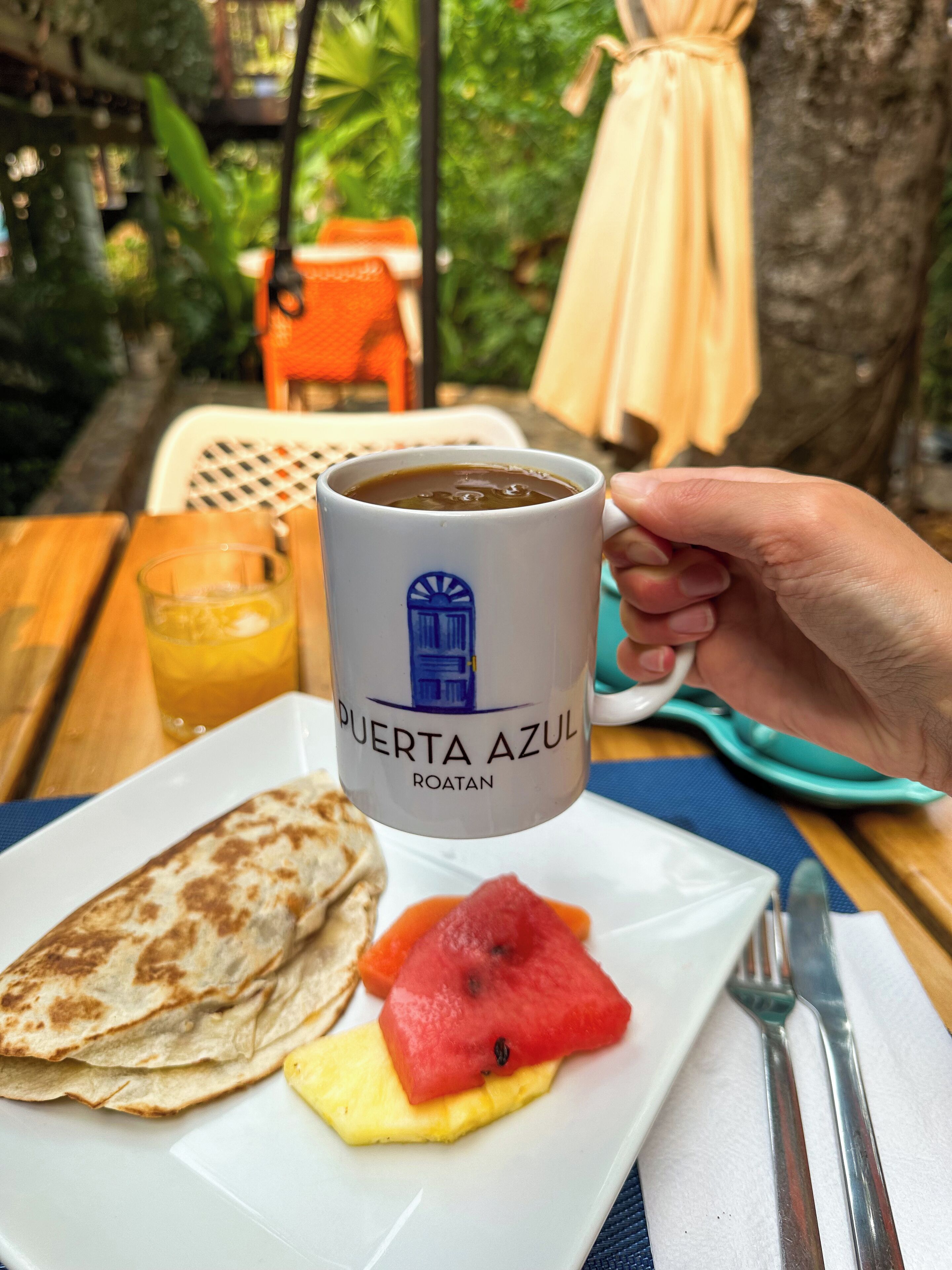 A hand holding a coffee mug labeled "PUERTA AZUL ROATAN" with a plate of food including a quesadilla, watermelon, and pineapple.