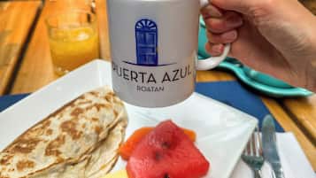 A hand holding a coffee mug labeled "PUERTA AZUL ROATAN" with a plate of food including a quesadilla, watermelon, and pineapple.