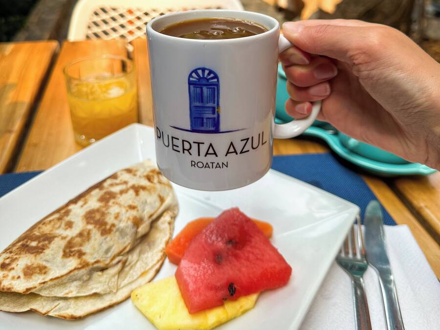 A hand holding a coffee mug labeled "PUERTA AZUL ROATAN" with a plate of food including a quesadilla, watermelon, and pineapple.