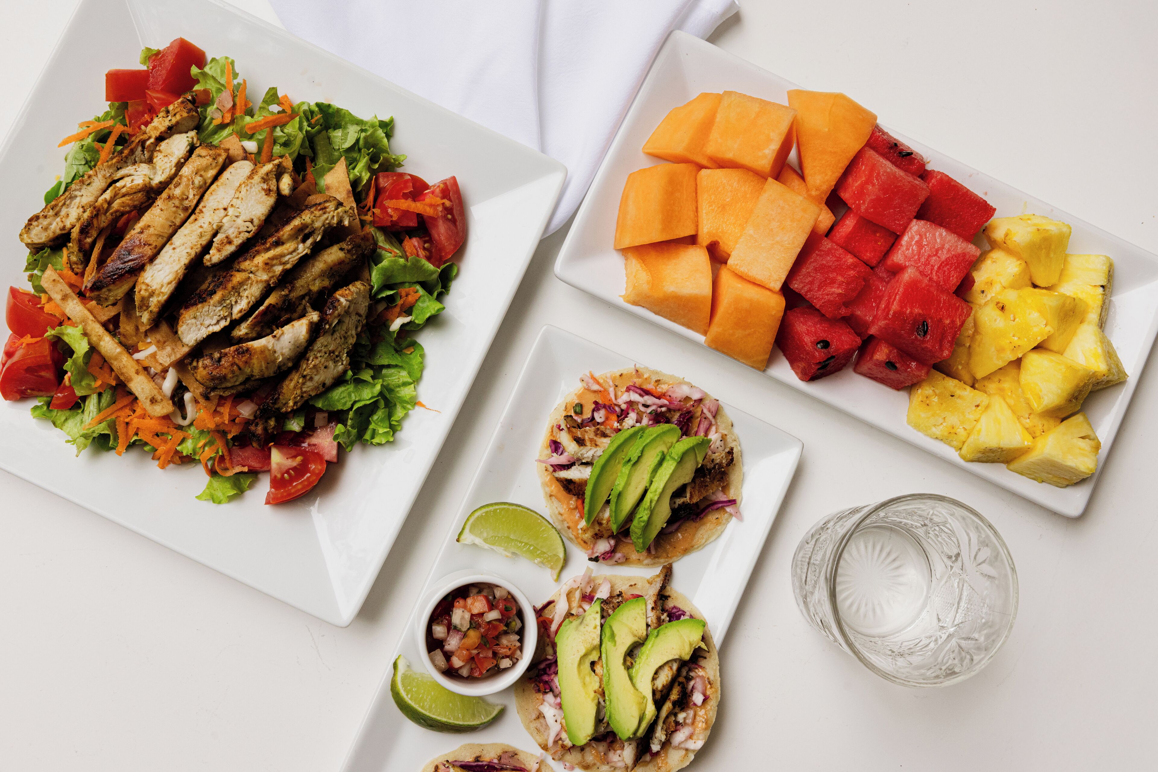 A table with various dishes including grilled chicken, a fruit platter, and tacos.