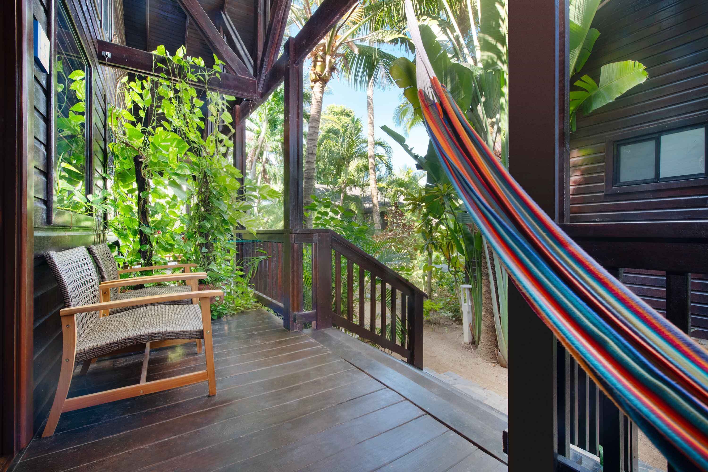 Una terraza de madera con una silla de mimbre, una hamaca y abundante vegetación tropical.