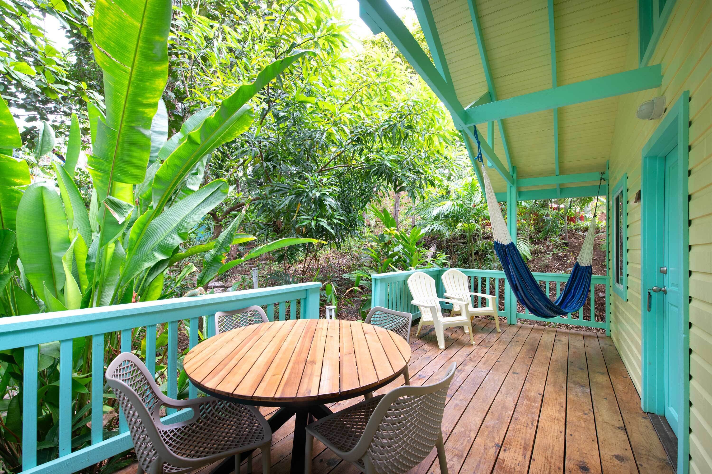 A wooden deck with a round table and wicker chairs, surrounded by lush greenery and a turquoise railing.