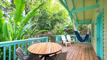 A wooden deck with a round table and wicker chairs, surrounded by lush greenery and a turquoise railing.