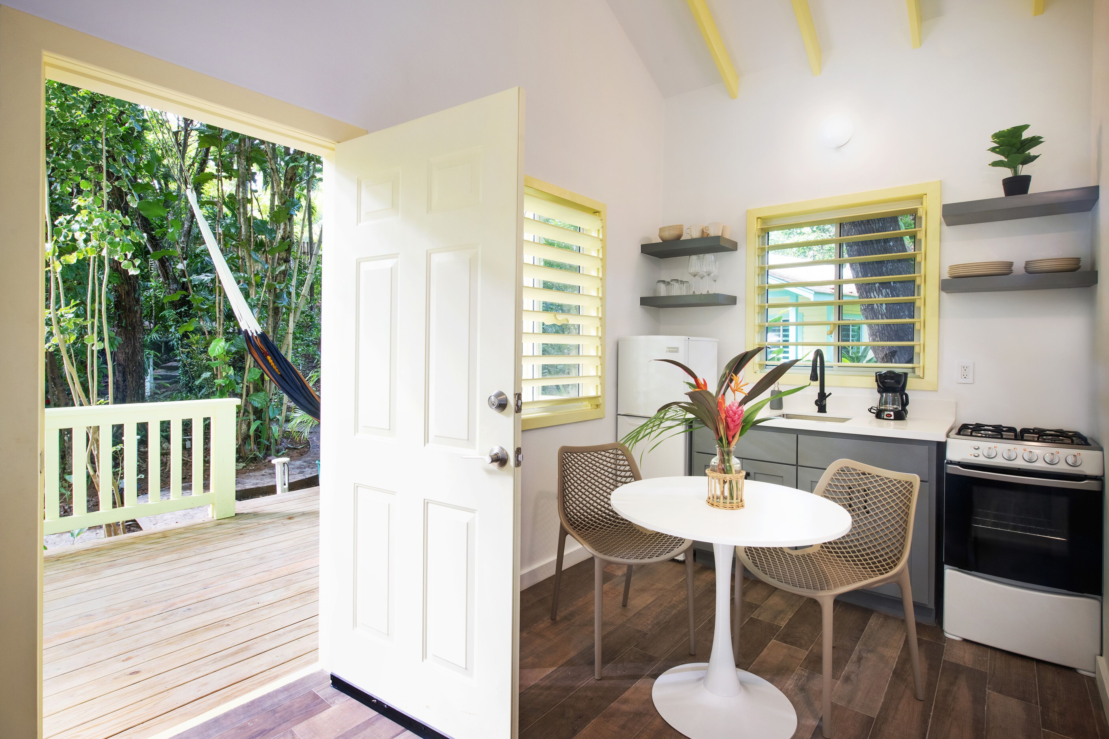 Una cocina con comedor, una puerta blanca que da a un balcón y vistas al exterior con vegetación.