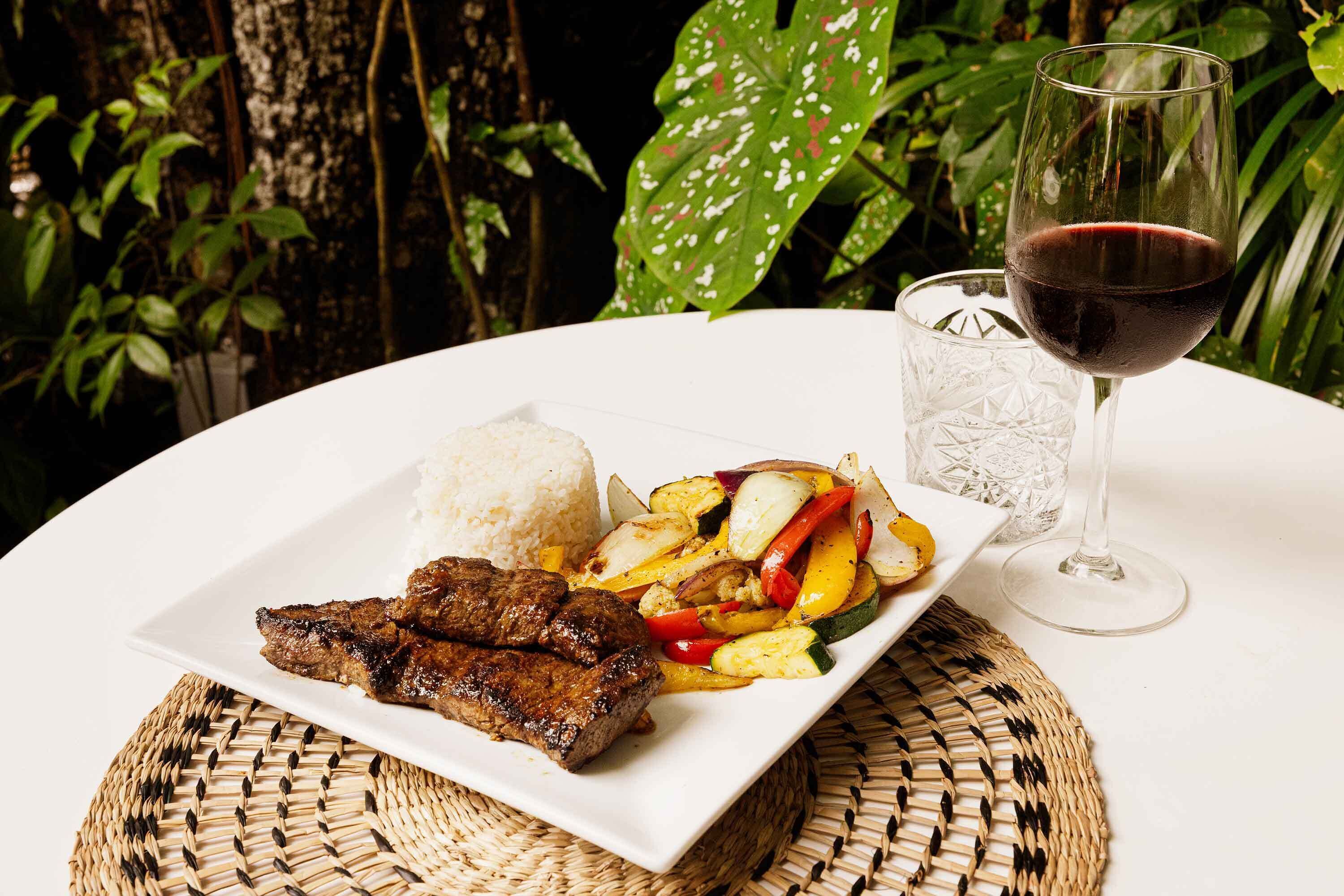 A plate of grilled meat, rice, and grilled vegetables on a white table with a glass of red wine.