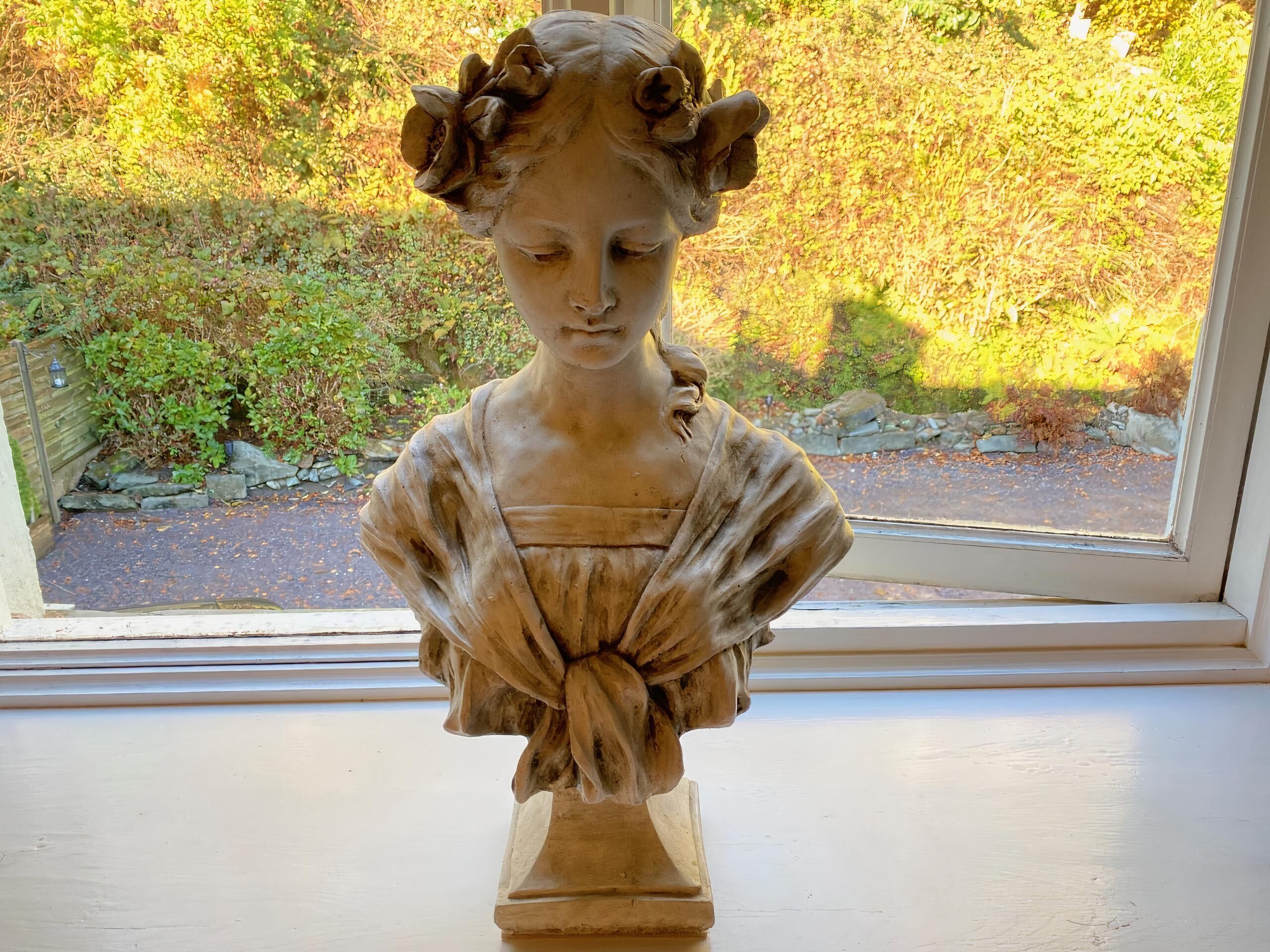 A bust sculpture on a pedestal by a window overlooking a garden.