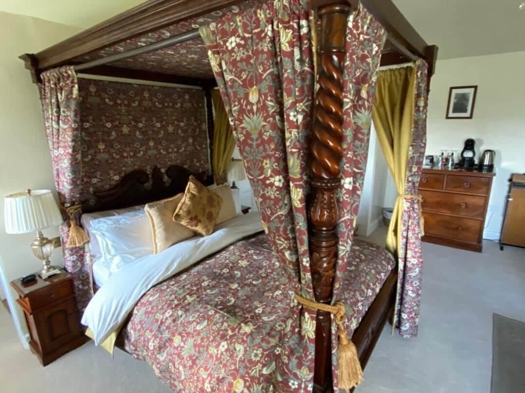 A four-poster canopy bed with floral bedding, a wooden nightstand with a lamp, and a dresser with a mirror.