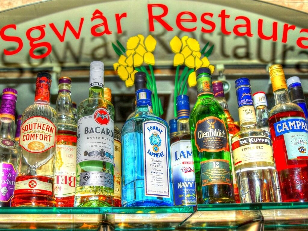 A variety of liquor bottles on a shelf with a sign reading "Sgwar Restaurans" in the background.