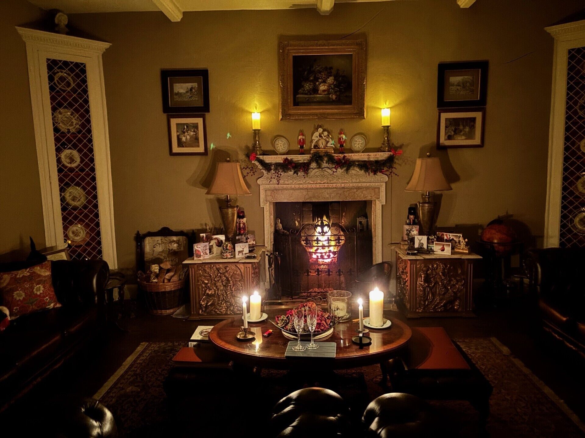 Une pièce chaleureuse, éclairée d’une lumière douce, avec une cheminée, des tableaux encadrés aux murs et une table décorée de bougies et de verres.