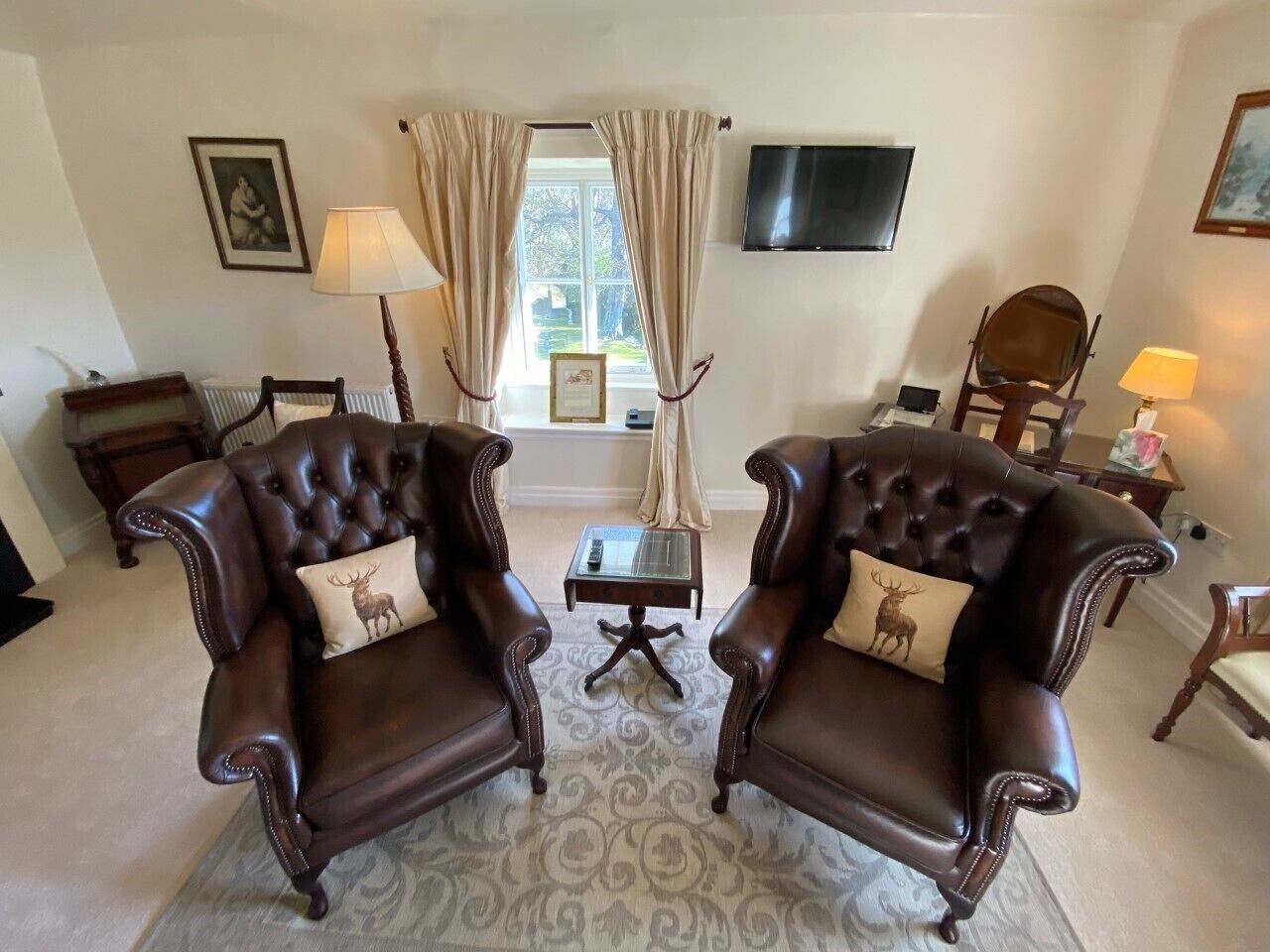 A living room with two leather armchairs, a glass coffee table, a television mounted on the wall, and a window with curtains.