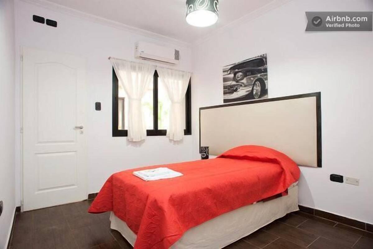 A single bed with a red bedspread, a white door, a window with white curtains, and a wall-mounted air conditioning unit.