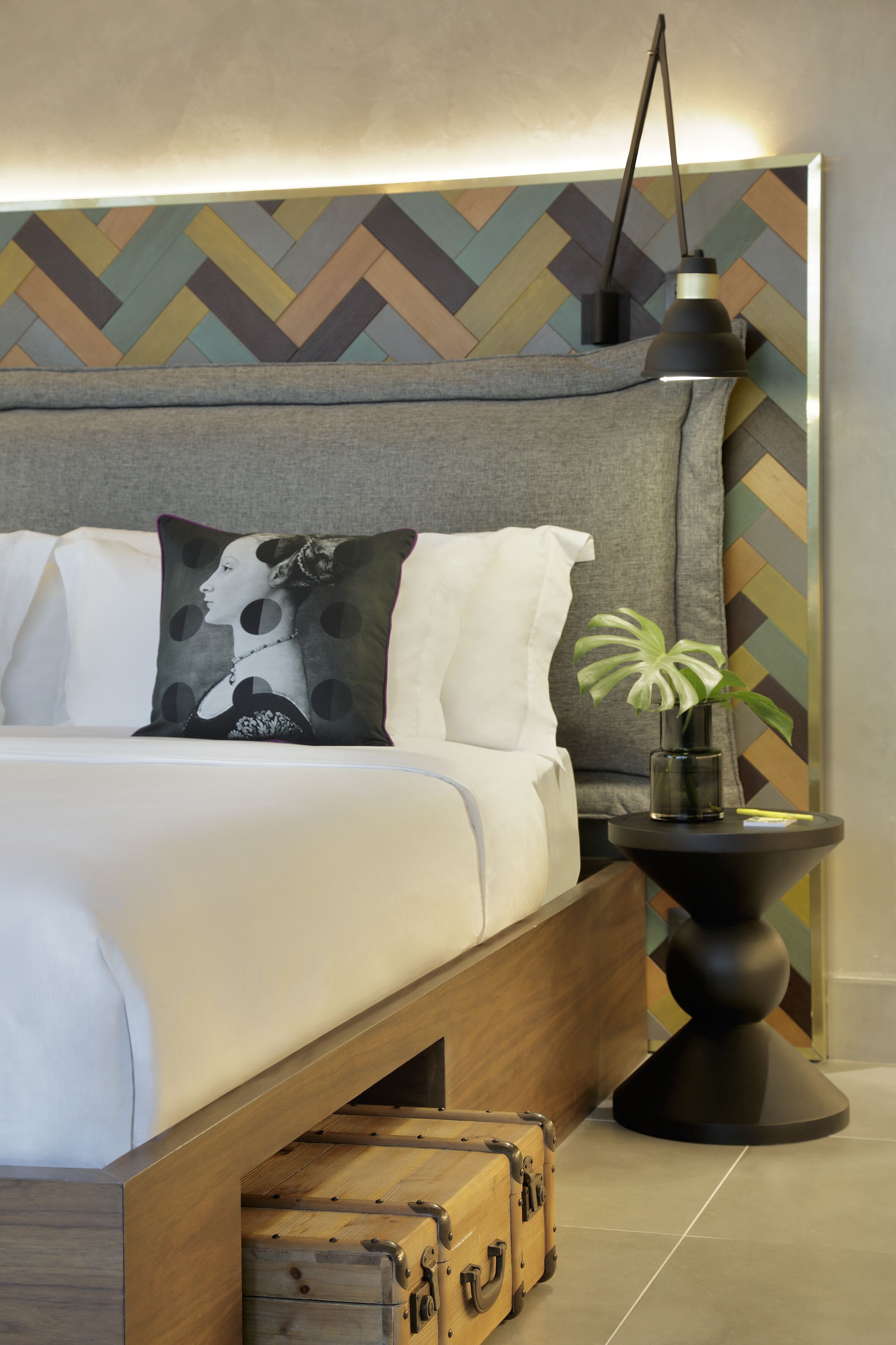 A modern bedroom with a bed, pillows, a bedside table with a lamp, a plant, and a vintage suitcase.