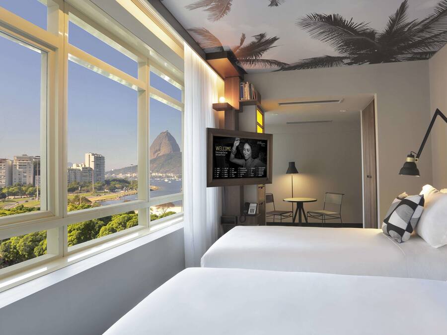 Habitación de hotel con un ventanal grande que ofrece vistas a la ciudad, una cama con almohadas, un escritorio con lámpara y un televisor en la pared.