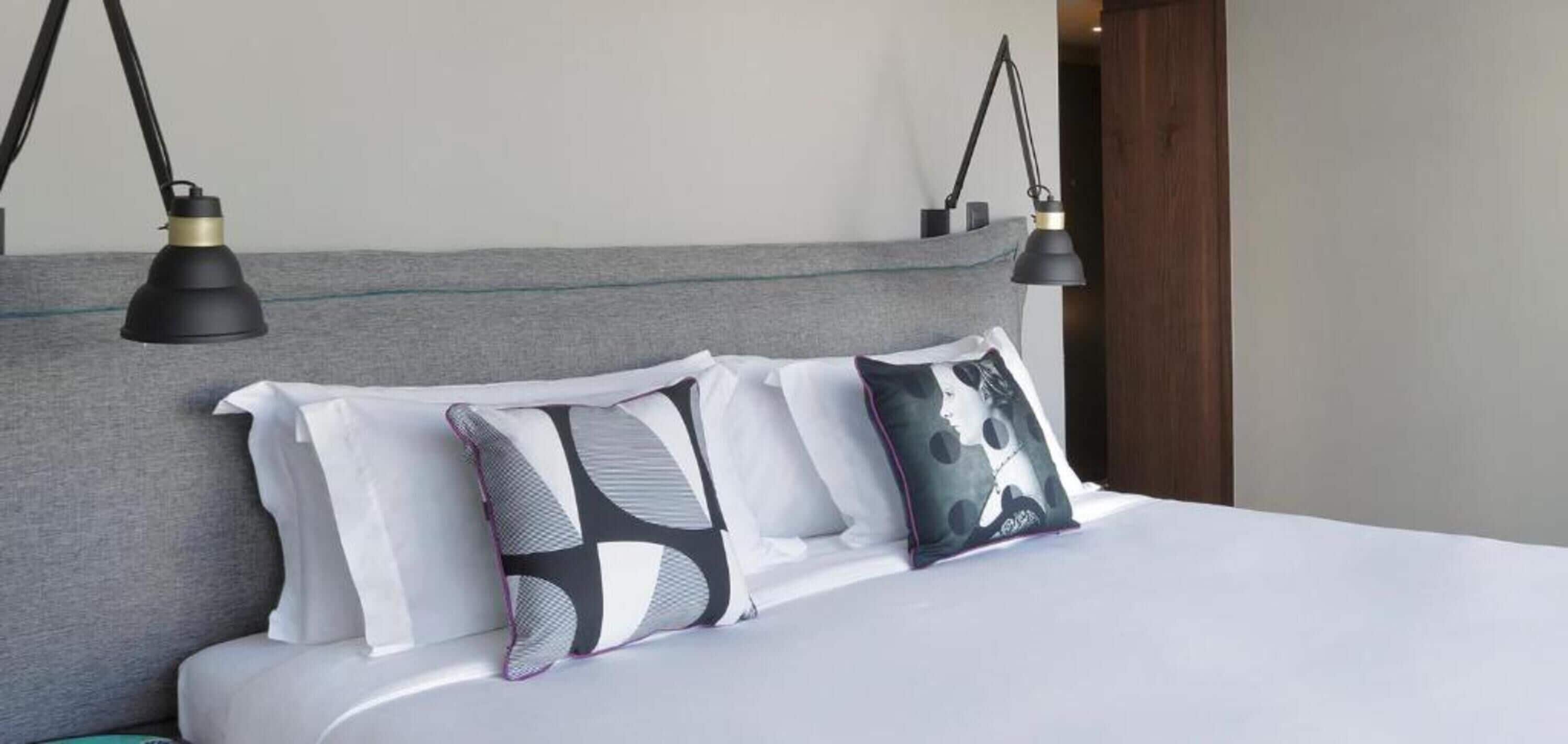 Un lit avec une literie blanche, deux lampes noires et trois coussins à motifs.