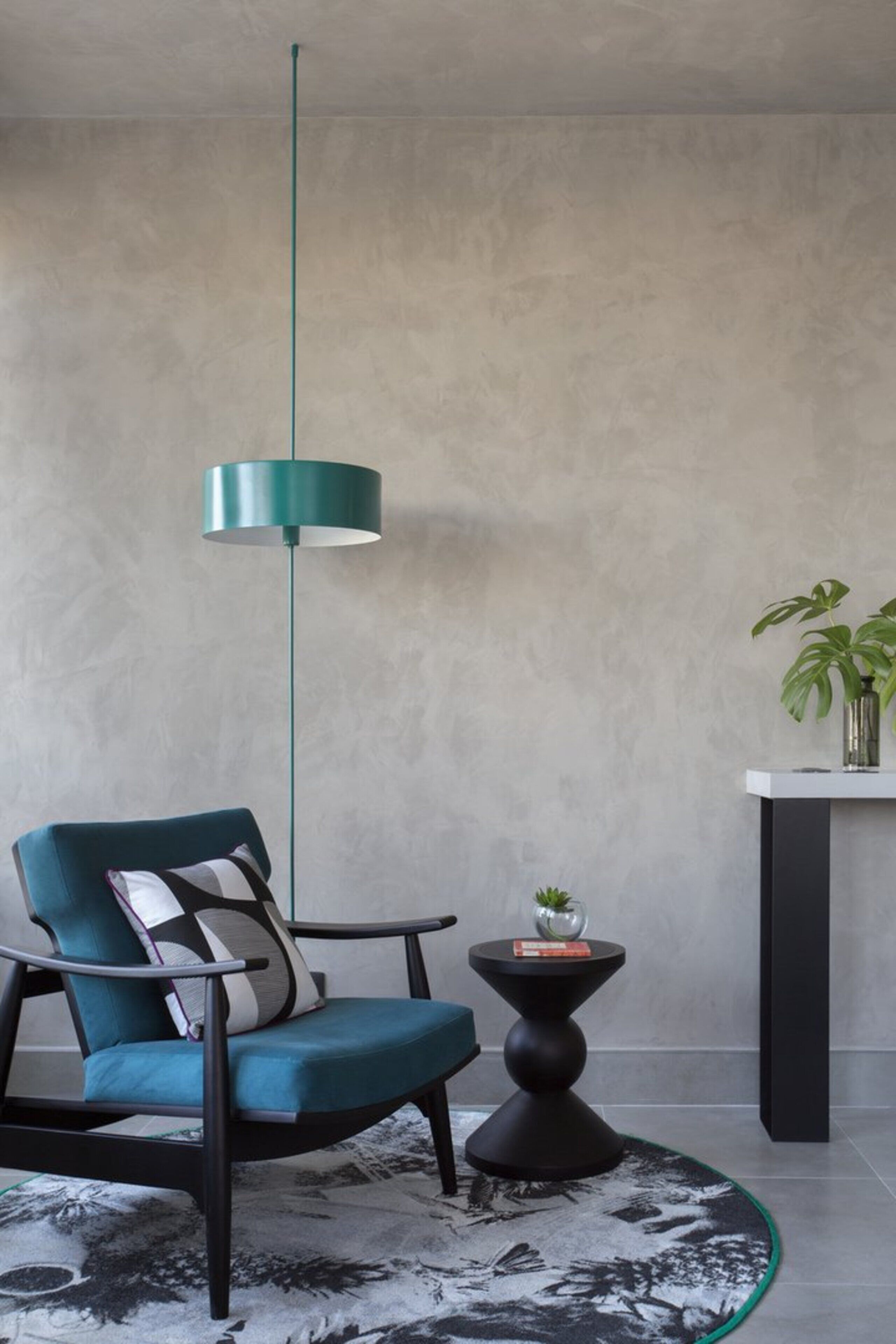 A modern room with a teal chair, a black and white rug, a round black table with a plant, and a teal pendant light.