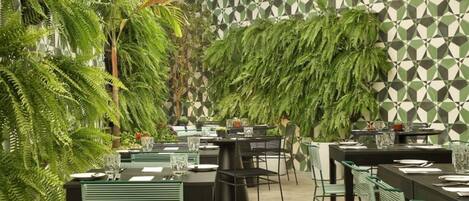 An indoor dining area with multiple tables and chairs, surrounded by lush green plants and a patterned wall.
